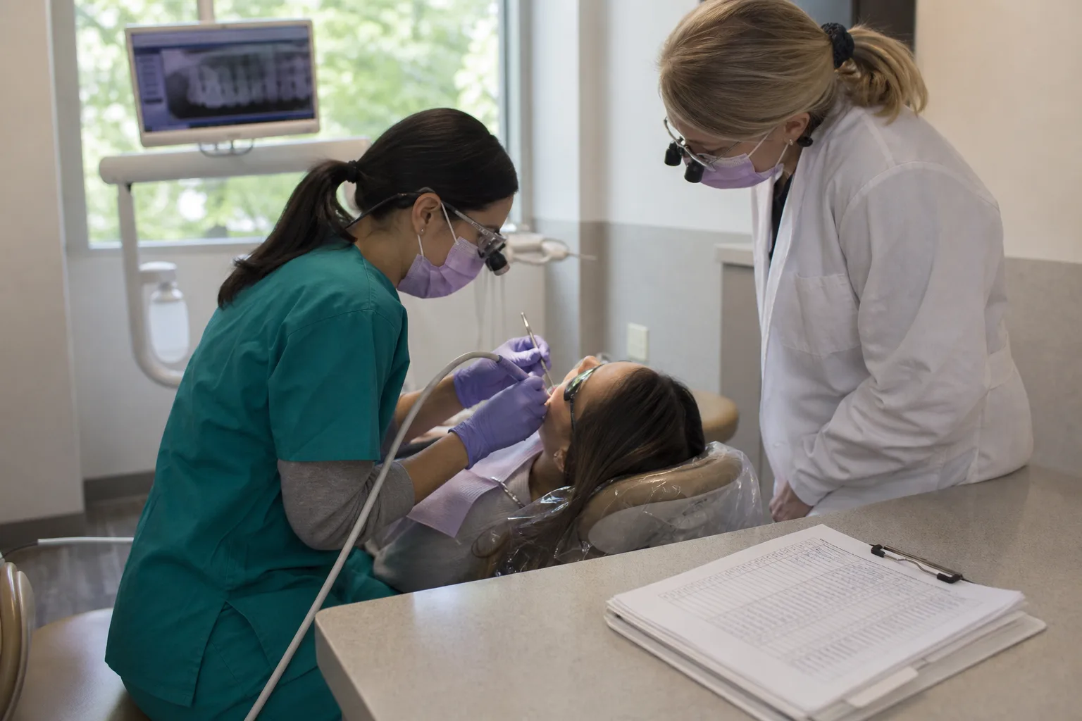 A dental hygiene student in scrubs working alongside a supervising clinician in a real clinic operatory, with a clipboard of signed hour logs on the counter