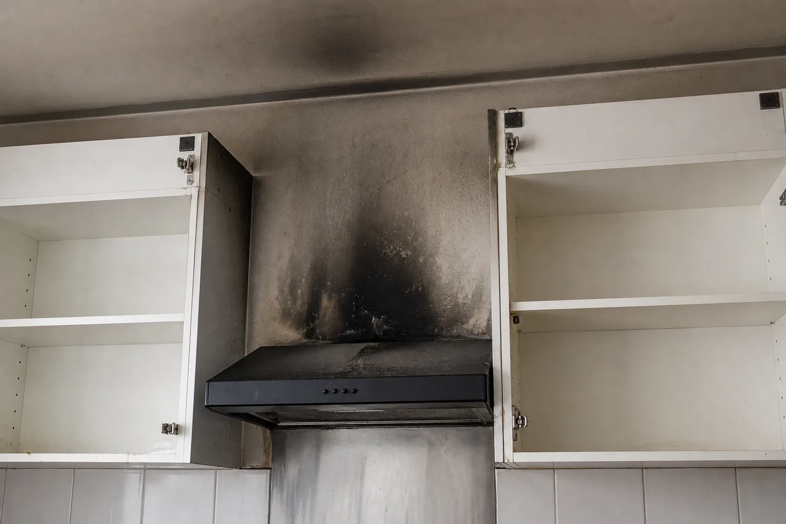 A kitchen wall with soot staining above a range hood and cabinet doors removed from their hinges
