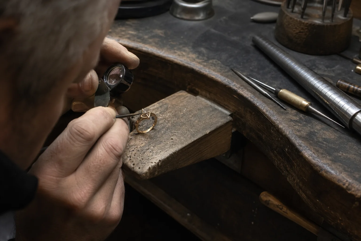 A jeweler's bench with a gold ring held in a bench block, worn prongs visible under a loupe, small hand tools and a ring mandrel beside it