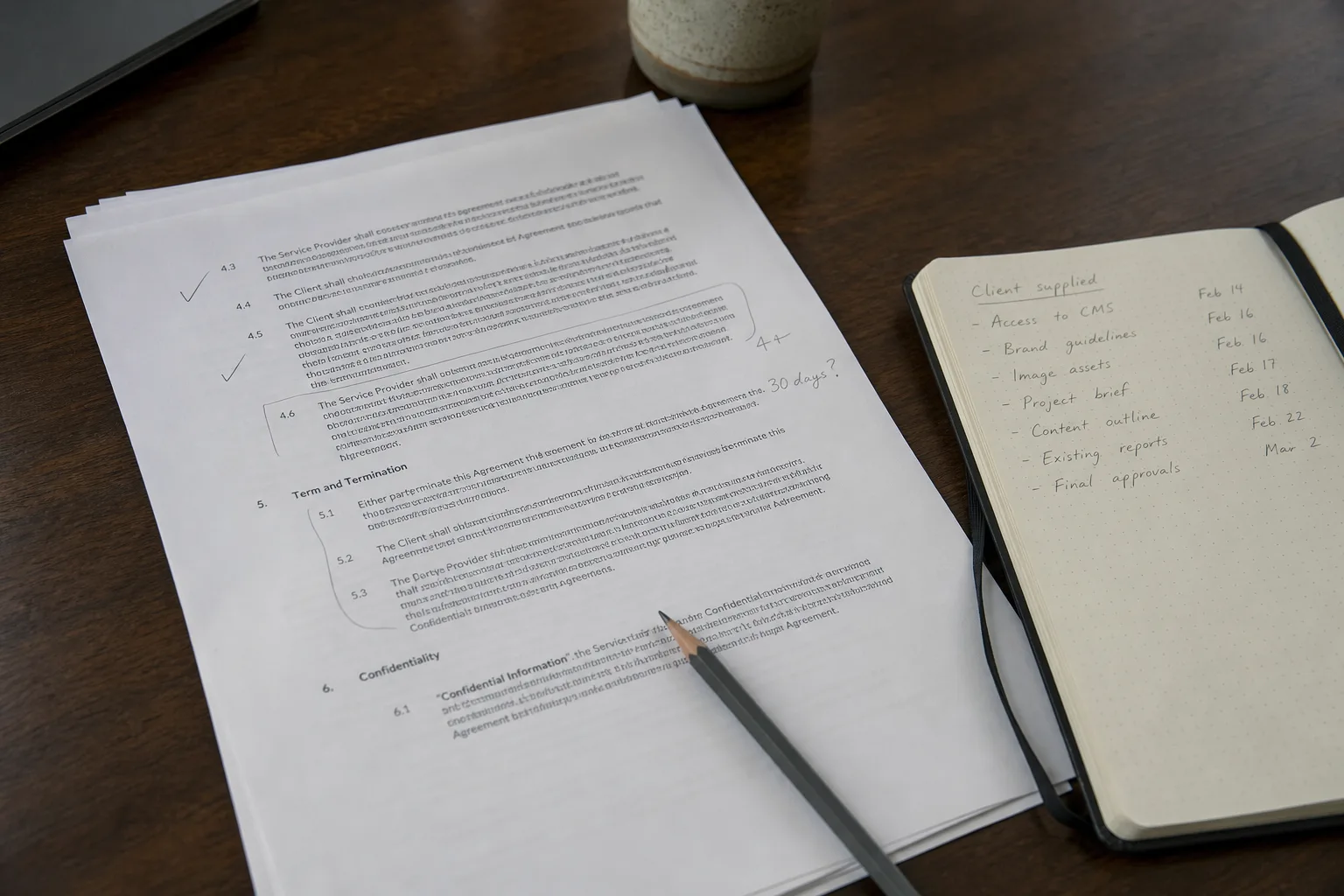 A printed multi-page services contract on a desk with several clauses marked in pencil, beside a notebook listing client-supplied items and dates