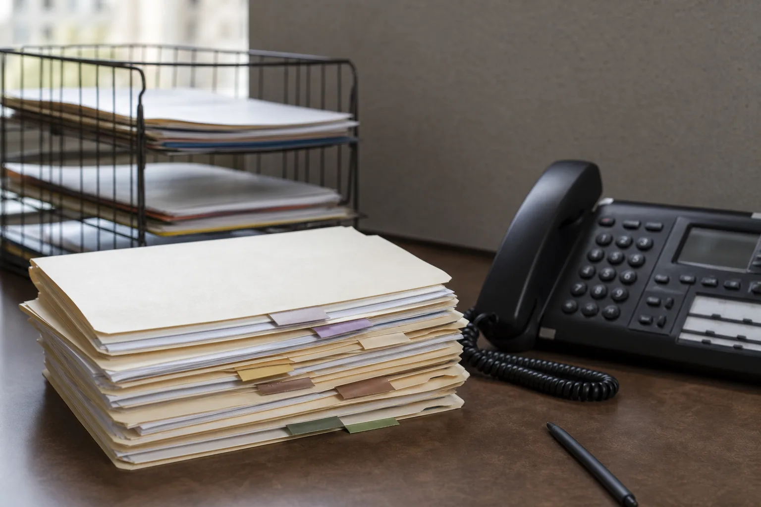 A stack of labeled file folders on a desk beside a telephone and a wire document tray